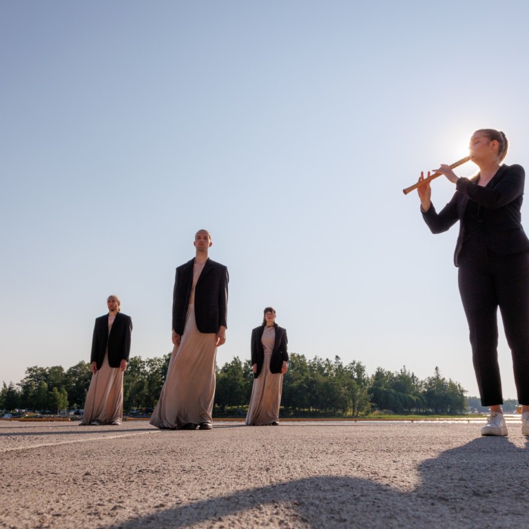 A woman playing a flute and three dancers standing behind in dresses