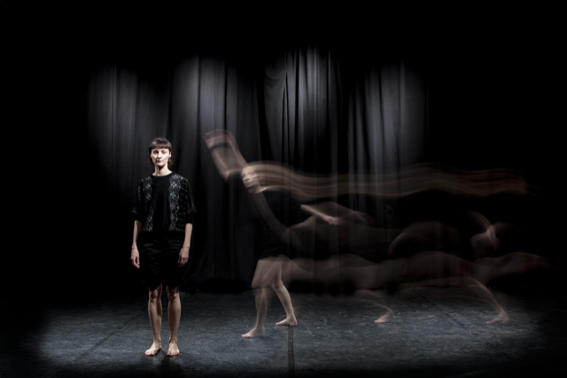 A woman stands on a black stage in black clotihng