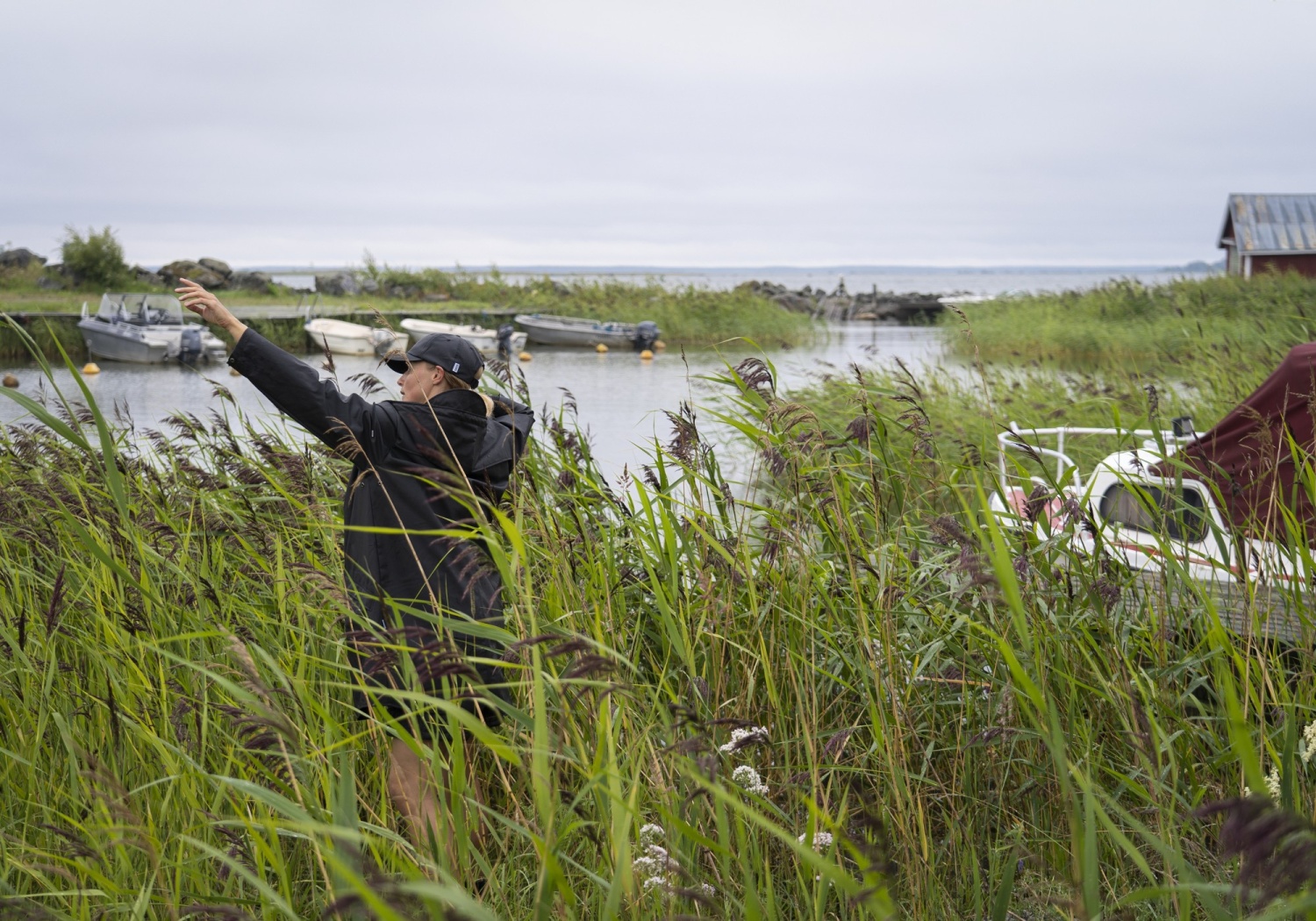 Nainen tanssii mustissa vaatteissa meren äärellä kaislikossa.
